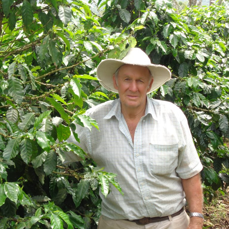 Carlos Fernandez Morera wearing a wide-brimmed light beige hat, a light-colored short-sleeve button-up shirt, and khaki pants stands in front of dense green coffee plants at Finca El Diamante Costa Rica.