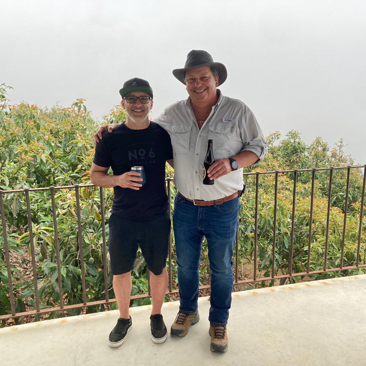 Denis from No6 Coffee and Ricardo Zelaya standing together on a concrete balcony with a metal railing and green foliage in the background under an overcast sky. The man on the right wears a cowboy hat, a light gray button-up shirt, blue jeans, and brown boots, holding a dark bottle.
