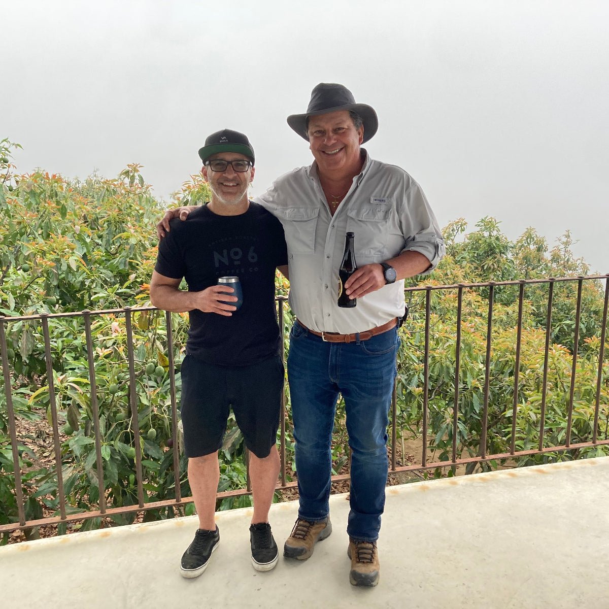 Denis from No6 Coffee and Ricardo  Zelaya standing together on a concrete balcony with a metal railing and green foliage in the background under an overcast sky. The man on the right wears a cowboy hat, a light gray button-up shirt, blue jeans, and brown boots, holding a dark bottle.